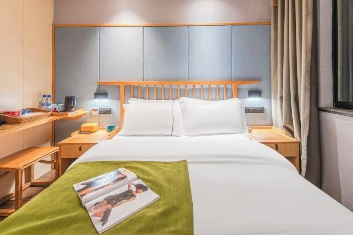 A hotel room with a bed, bedside tables, a nightstand, a lamp, a window with curtains, and a magazine on the bed.