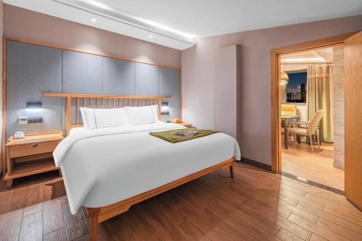 A hotel room with a large bed, wooden headboard, bedside tables, and a view of the city through an open door.