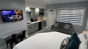 Ett modernt och kompakt hotellrum med ett litet pentry, en platt-TV som är monterad på väggen, en säng med vita sängkläder, en grå soffa med kuddar och ett fönster med persienner.
