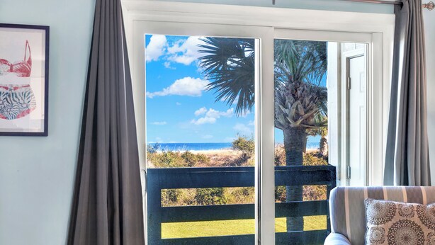 Un balcon avec vue sur la plage et les palmiers, une œuvre d’art encadrée au mur et un canapé à rayures agrémenté d’un coussin à motifs.