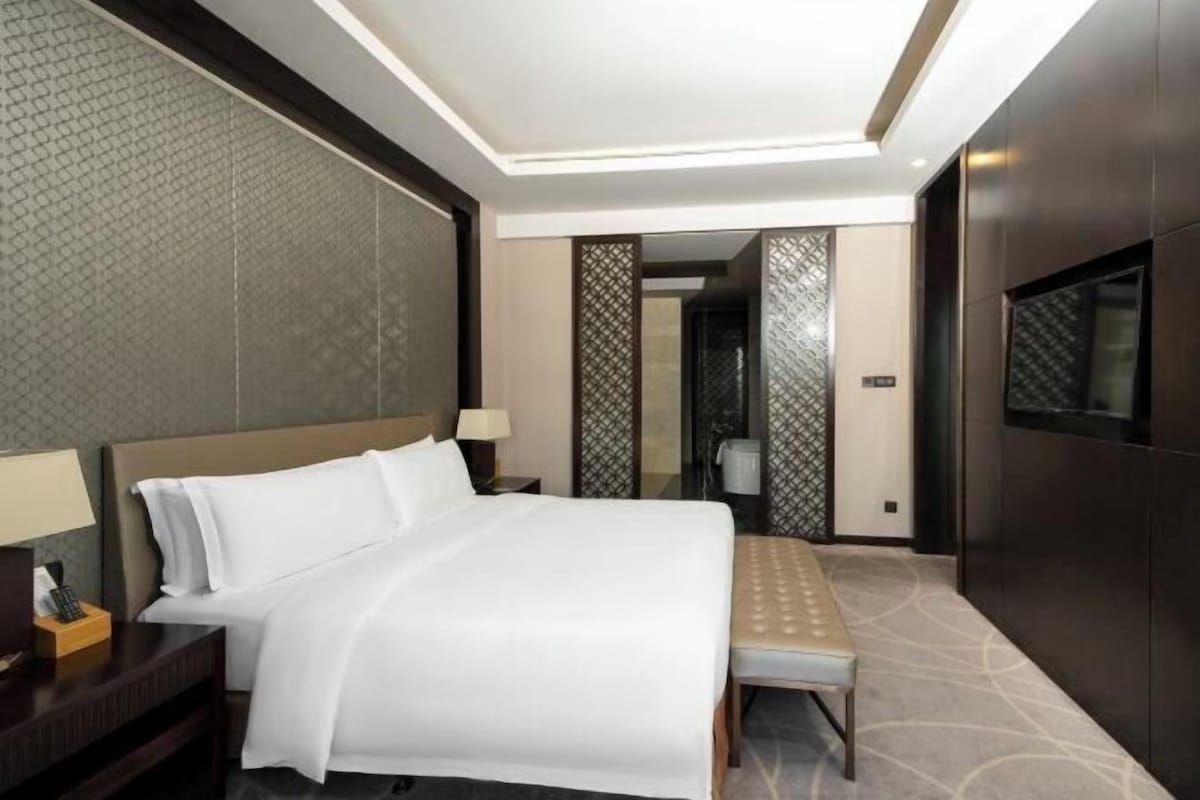 A hotel room with a large bed, bedside tables, a bench, a TV, and a patterned wall.