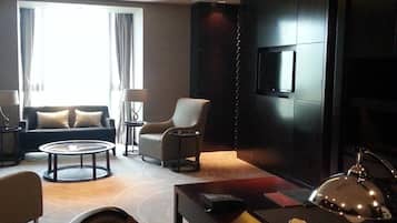 A modern hotel room with a sofa, armchairs, a coffee table, a TV, and a desk with a lamp.
