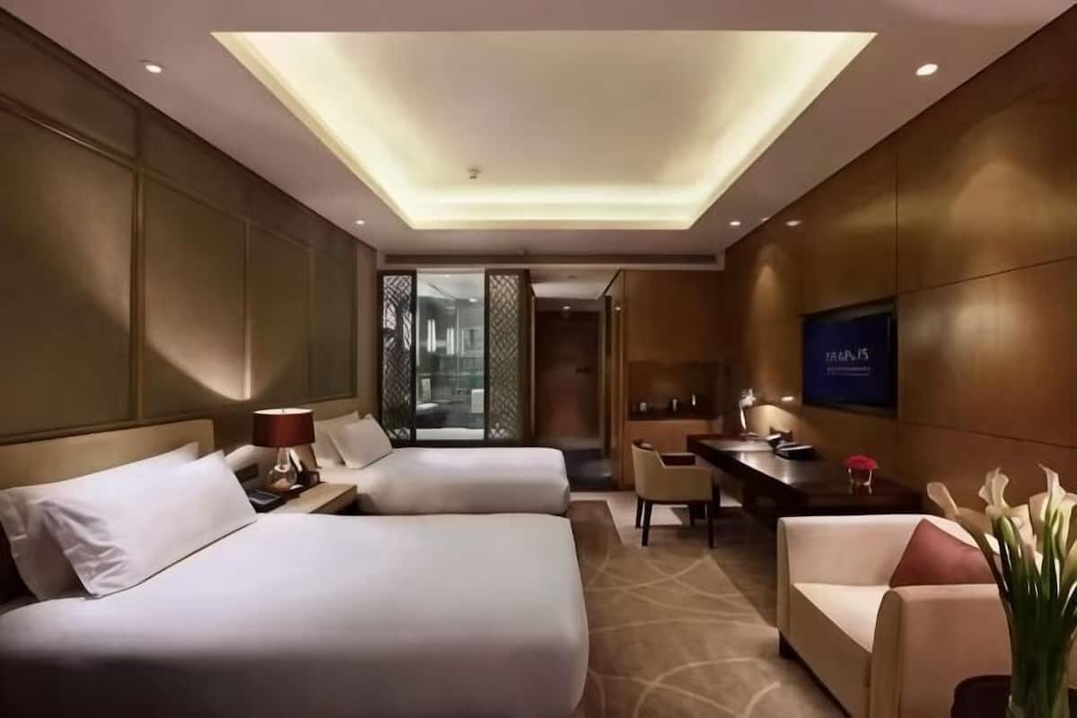 A hotel room with two beds, a desk, a chair, a TV, and a view of the outside.