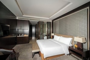 A hotel room with a bed, bedside table, lamp, TV, and a seating area with a bench.