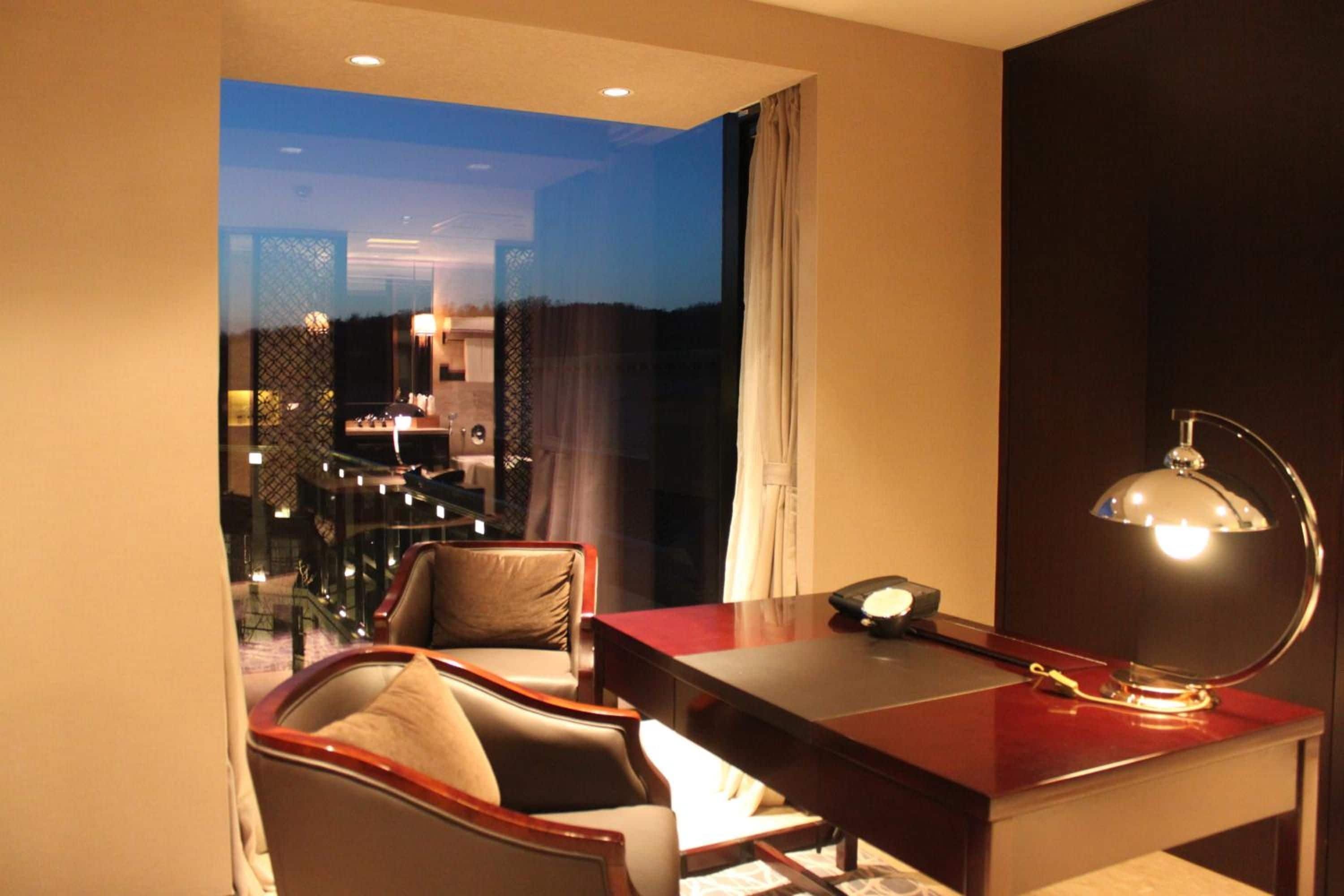 A hotel room with a desk, chair, and a view of the cityscape at dusk.