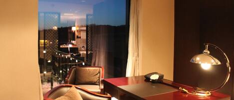 A hotel room with a desk, chair, and a view of the cityscape at dusk.