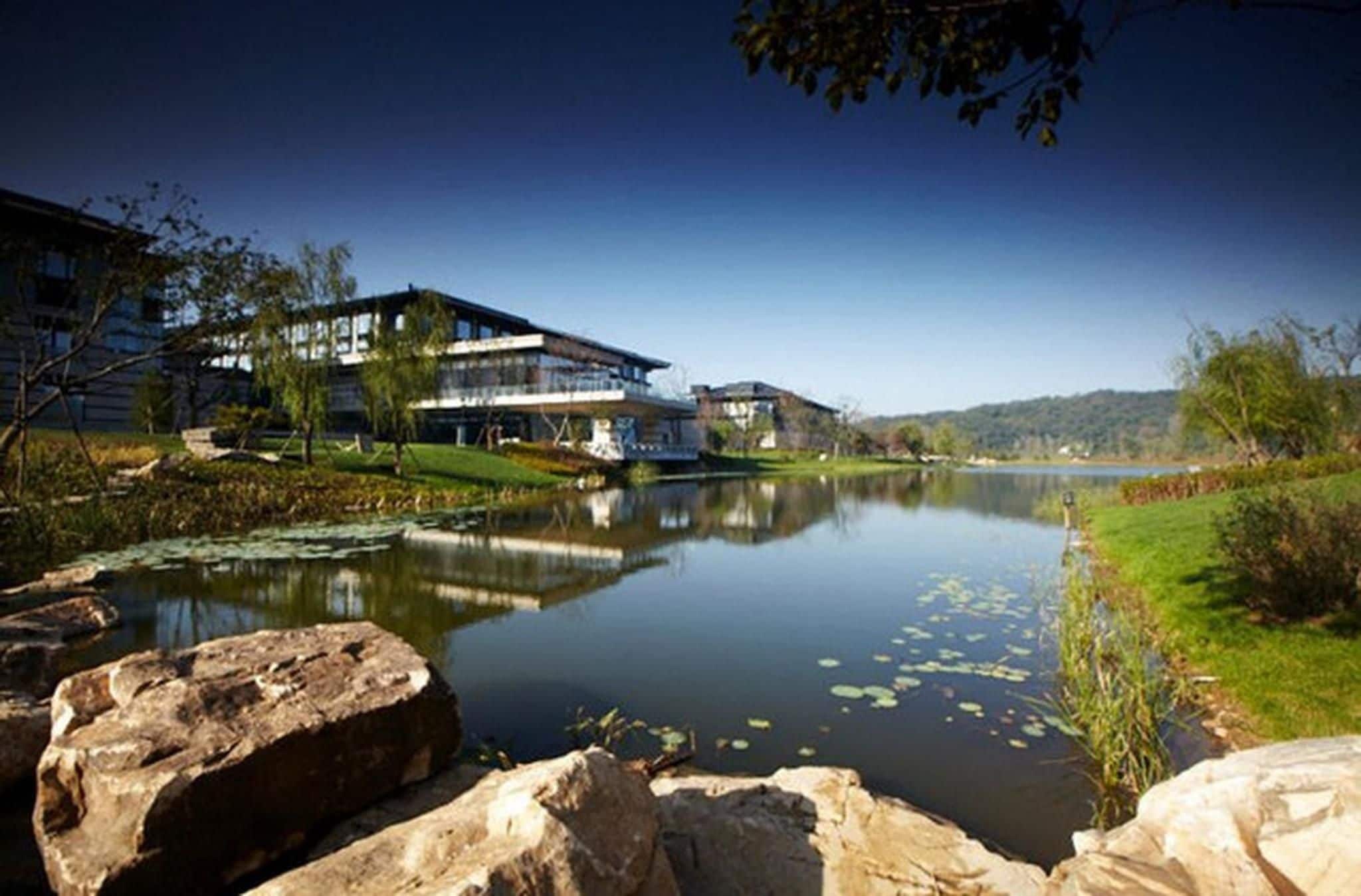 A lakeside view with modern buildings, a calm lake, and surrounding greenery.