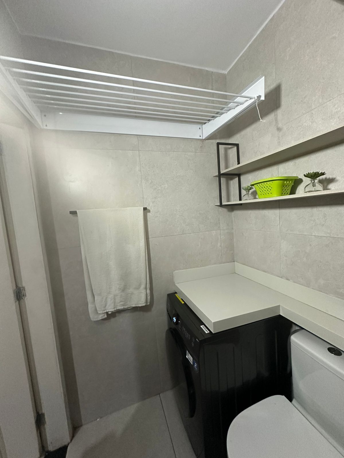 A compact bathroom with a white sink, a towel hanging on the wall, and a shelf with a green basket and a small plant.