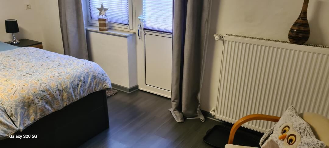 A bedroom with a bed, a chair, a radiator, a window with blinds, and a small table with a lamp.