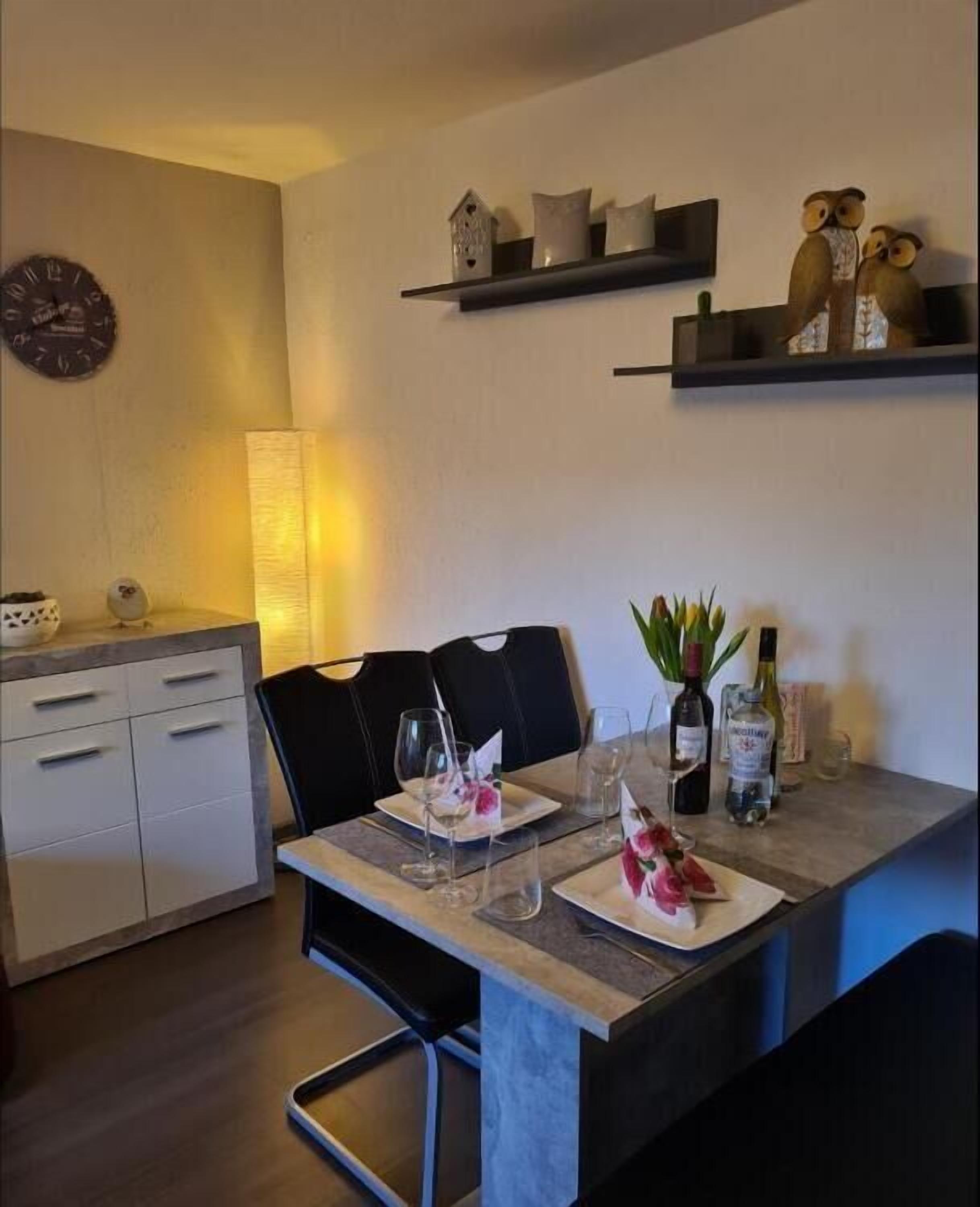 A dining area with a table set for two, a clock on the wall, and shelves with decorative items.