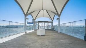 Una terraza abierta con baranda de cristal, una estructura central blanca y vistas despejadas del agua y el horizonte urbano distante.
