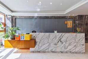 Ein Hotel-Rezeptionsbereich mit einer Marmor-Theke, einem Holzschrank mit Blumentöpfen, einer Wanduhr und einem Schwarzdbrett.