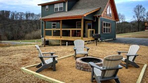 Una casa de dos pisos con un porche, una fogata y sillas Adirondack.