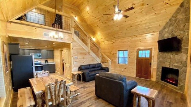 Un interior espacioso de cabaña de troncos con comedor, sofá, chimenea, cocina y una escalera que sube a un nivel superior.