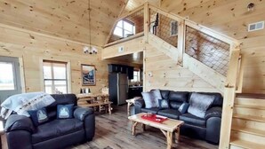 Interior de una cabaña de madera con un sofá de cuero negro, una mesa de centro de madera y una escalera a un nivel superior.