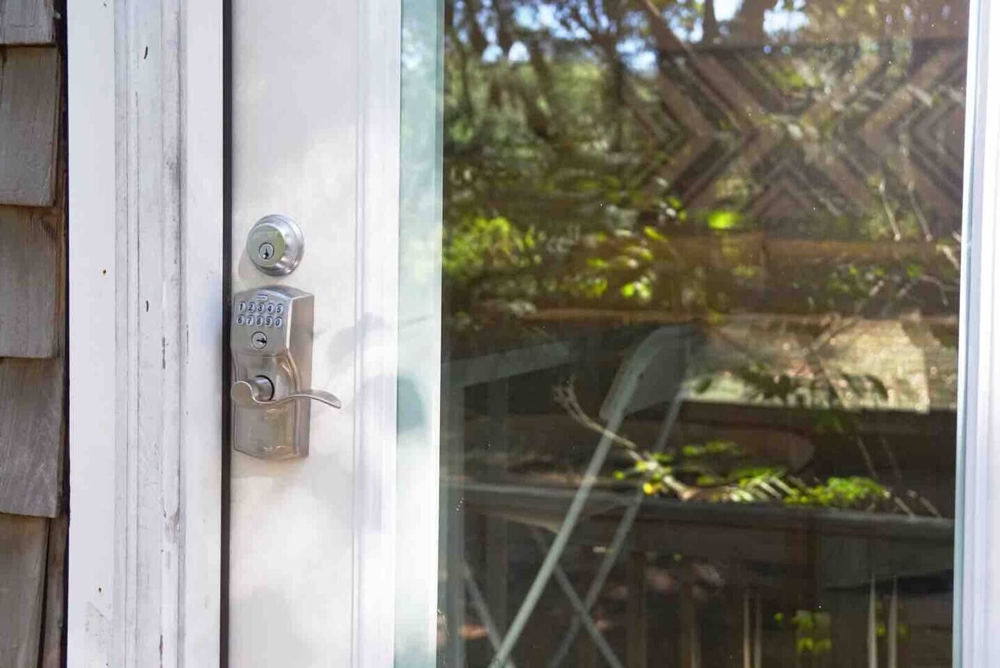 A door with a numeric keypad lock, a window, and a view of a wooden deck and greenery.