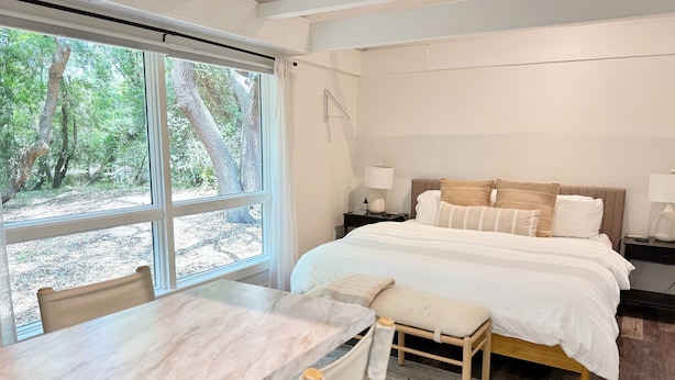 A bedroom with a bed, bedside tables, a chair, and a view of trees through large windows. - Live Oak Cottage - 'tiny house' / walk to beach (Kitty Hawk)