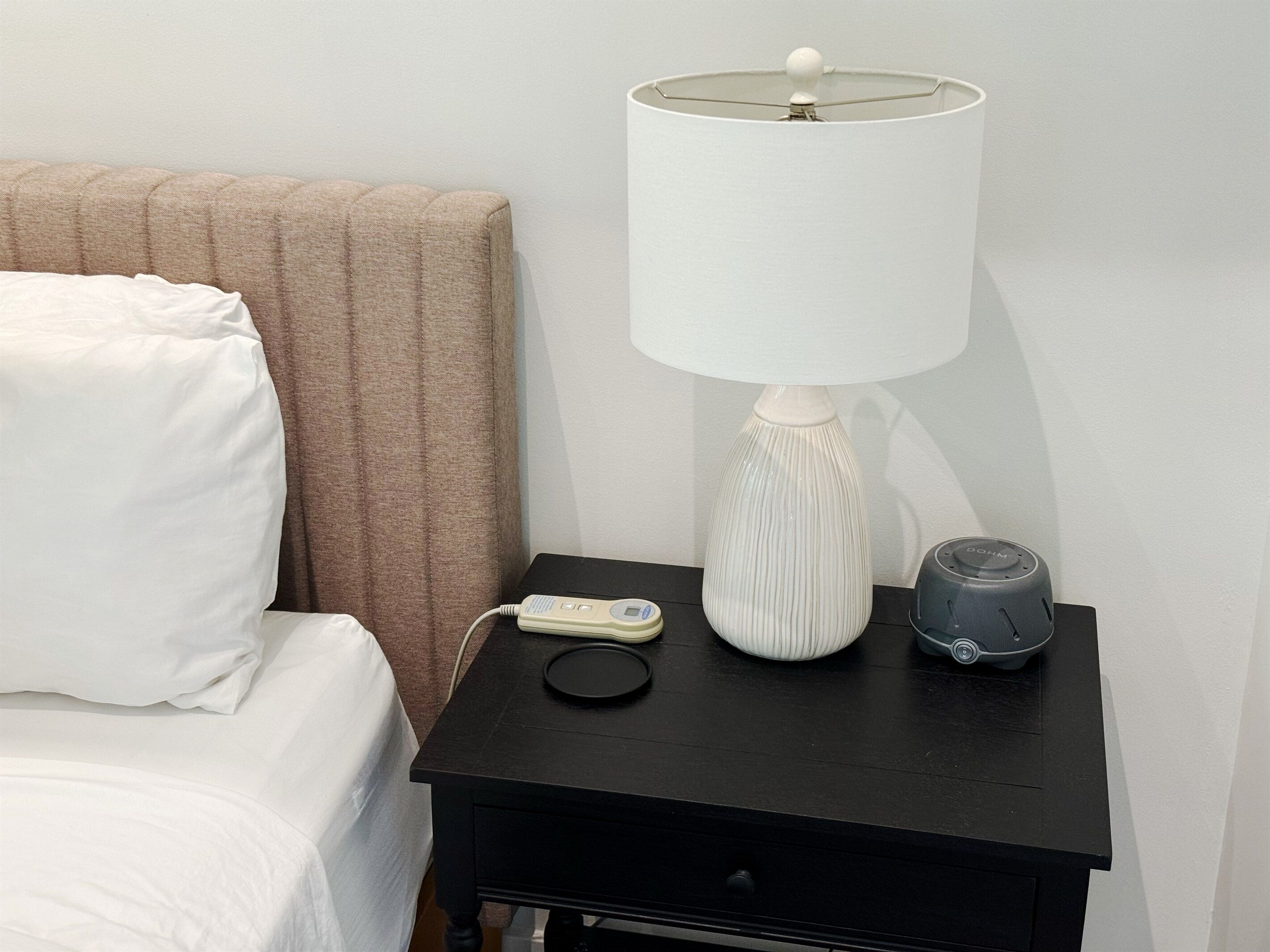 Een bed met witte lakens, een bruin hoofdeinde, een zwart nachtkastje met een witte lamp, een witte afstandsbediening en een grijze speaker.
