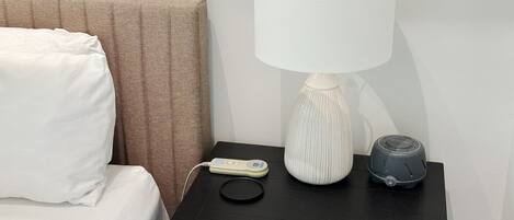 Een bed met witte lakens, een bruin hoofdeinde, een zwart nachtkastje met een witte lamp, een witte afstandsbediening en een grijze speaker.