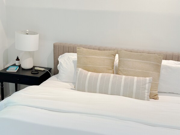 En pænt redt seng med hvide sengetøj og to beige puder. Et lille natbord med en hvid lampe, en bog og en flaske er synligt.