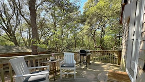 A wooden deck with white chairs and a grill, surrounded by trees. - Live Oak Cottage - 'tiny house' / walk to beach (Kitty Hawk)