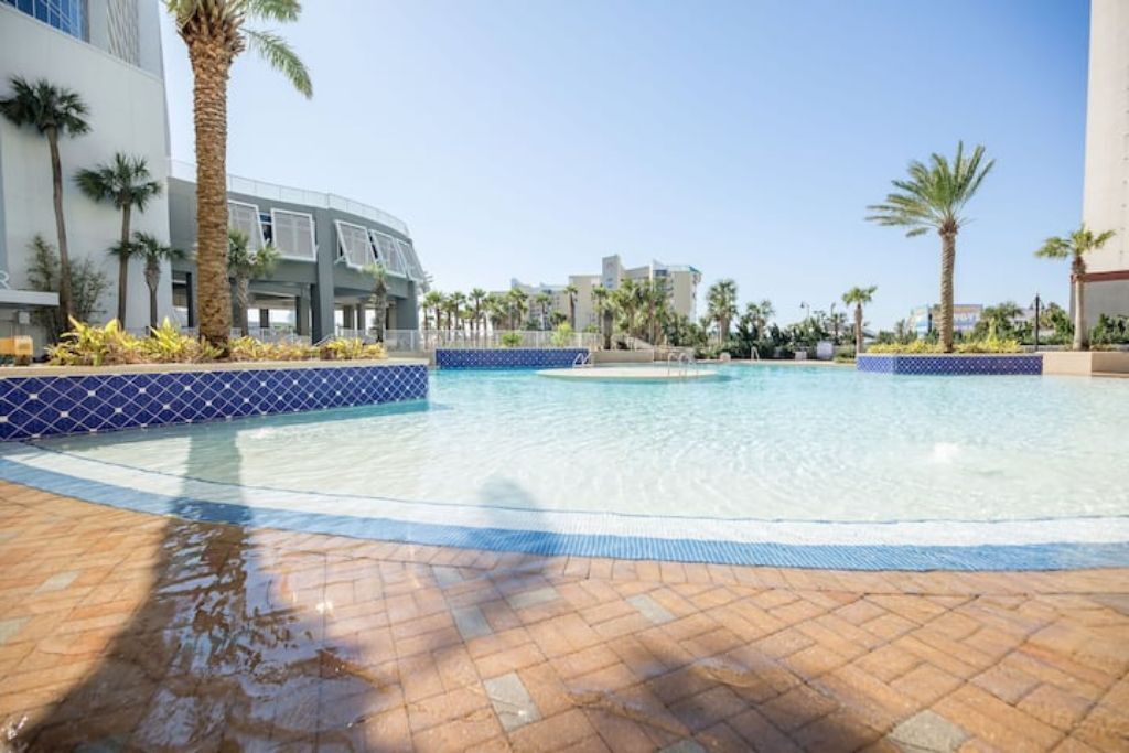 Une piscine bordée de carreaux, des palmiers et un bâtiment en arrière-plan.