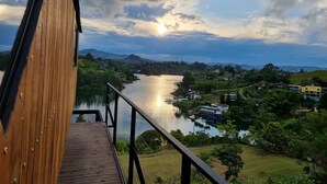 A wooden deck overlooking a river with houses and greenery in the background. - Woodside Ecoliving Guatape (Guatapé)