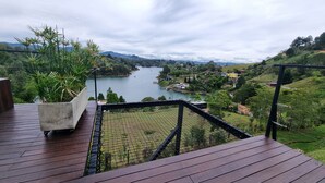 A wooden deck with a concrete planter of green plants, overlooking a river with boats, surrounded by hills and houses. - Woodside Ecoliving Guatape (Guatapé)