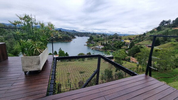 A wooden deck with a concrete planter of green plants, overlooking a river with boats, surrounded by hills and houses.