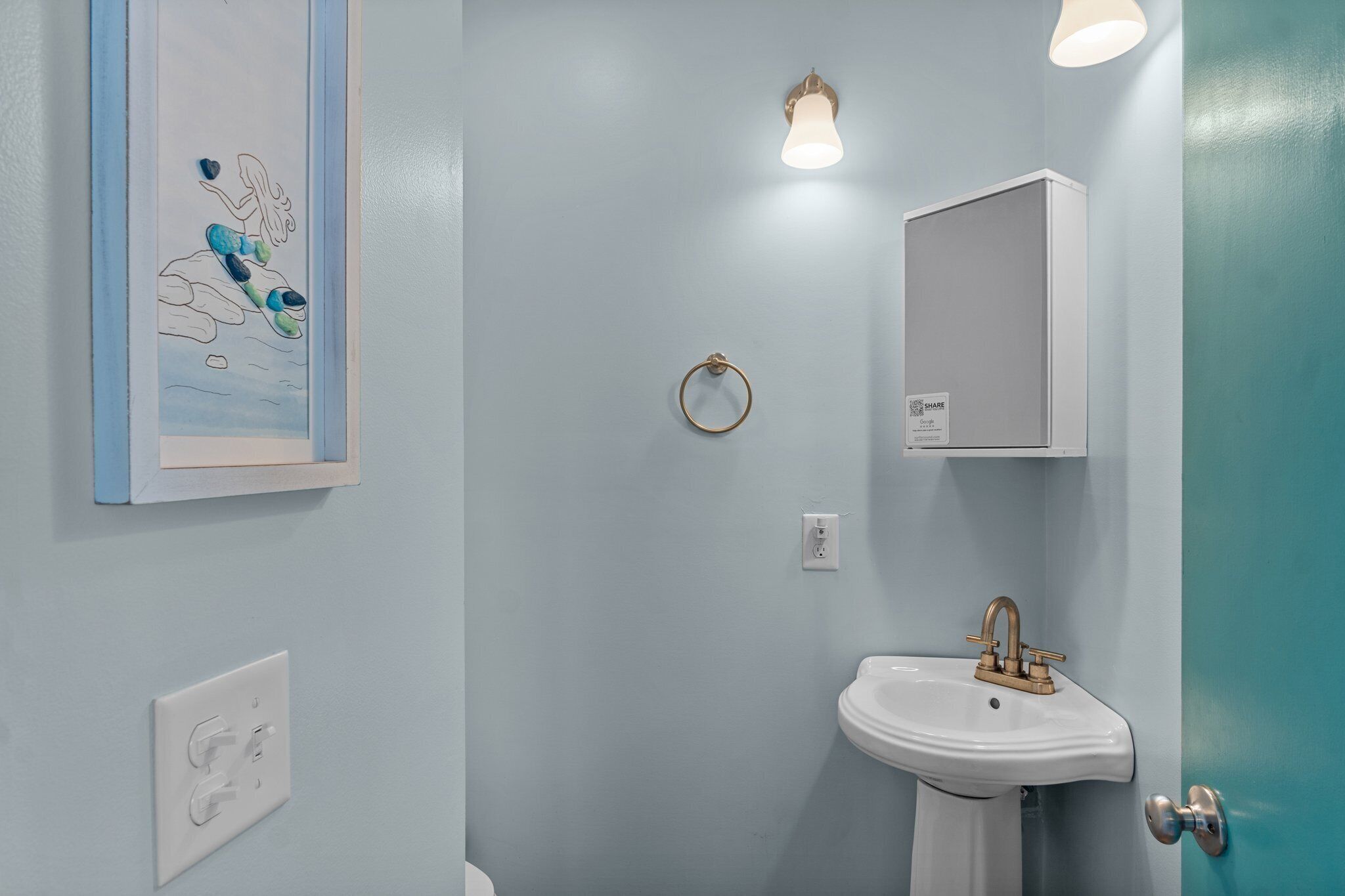 A bathroom with a light blue wall, a white sink with a gold faucet, a framed artwork, a towel ring, and a light fixture.