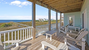 Una terraza de madera con barandillas blancas, un banco y sillas Adirondack con vistas a la playa.