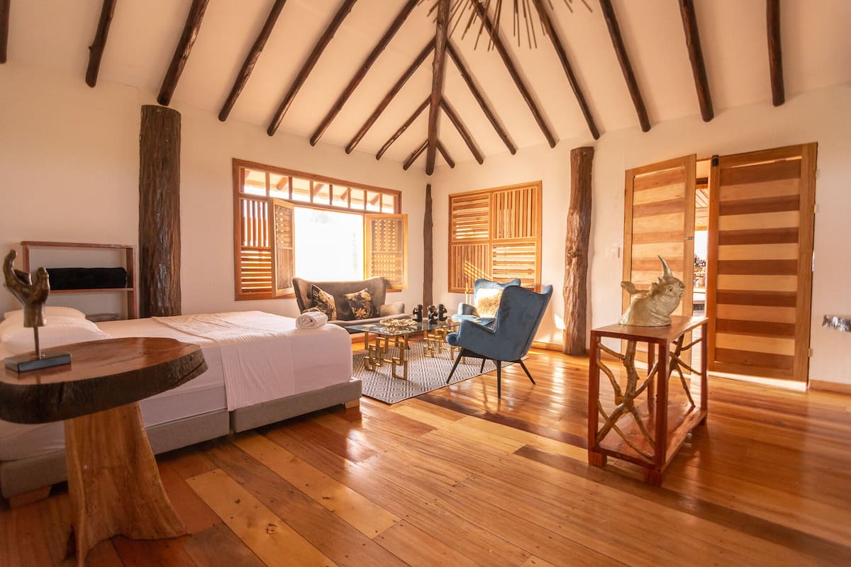 A spacious room with a high wooden ceiling, a bed, a blue armchair, and a small table with a sculpture.