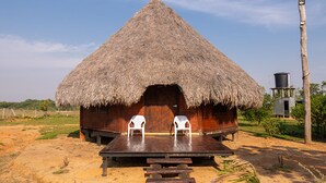 Una cabaña con techo de paja, dos sillas de plástico blancas en la parte delantera, una mesa de madera y una torre de agua al fondo.