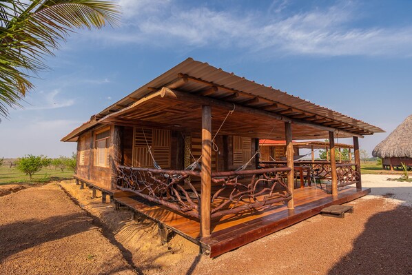 A rustic wooden cabin with a porch, surrounded by a sandy landscape and a palm tree.