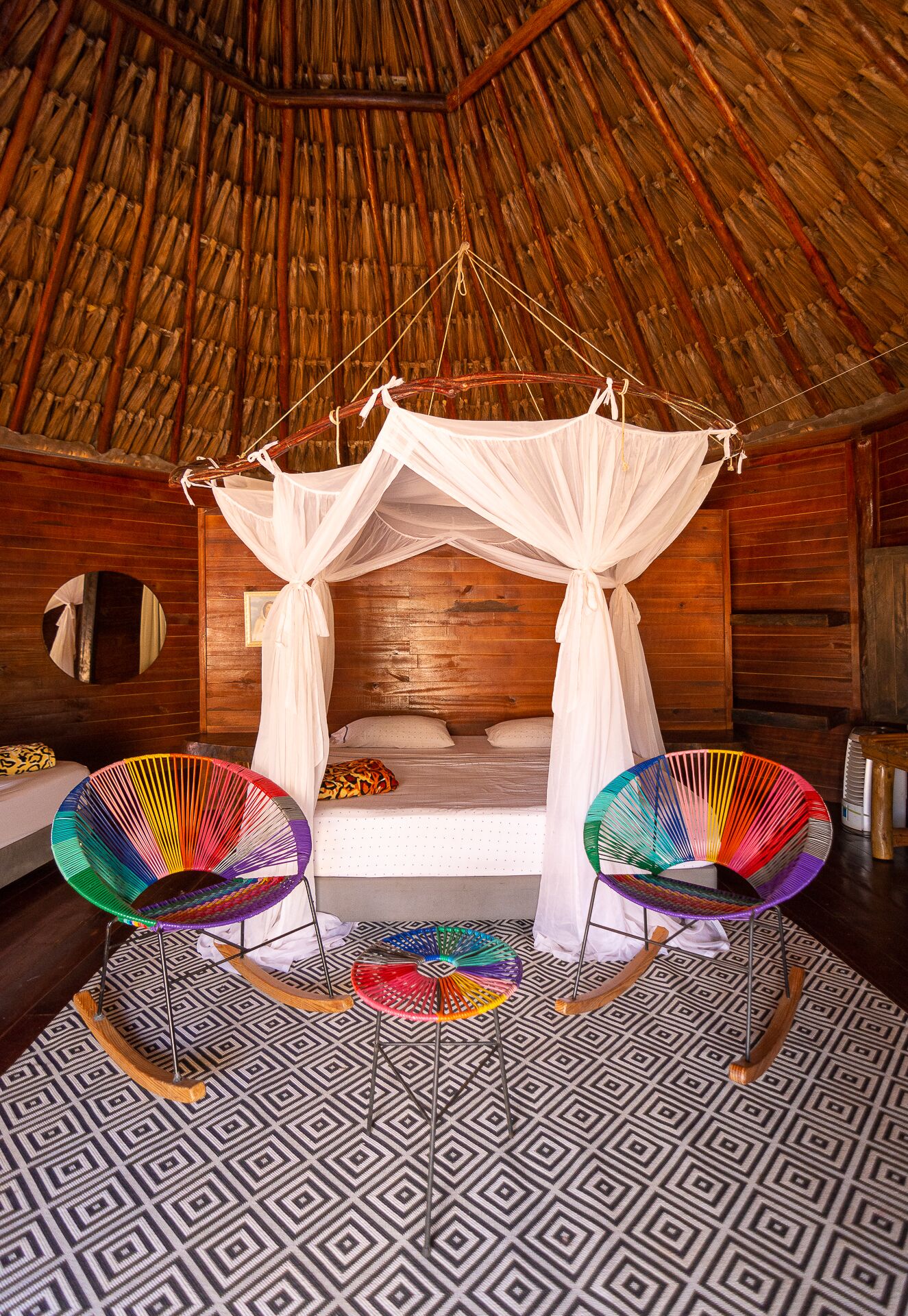 A room with a thatched roof, a bed with a canopy, and two colorful rocking chairs.