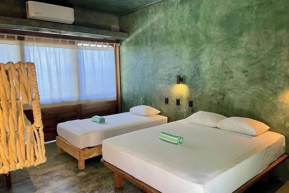 Ein Zimmer mit zwei Betten, einem Vorhangfenster, einer Wandleuchte und einem Korbsessel.