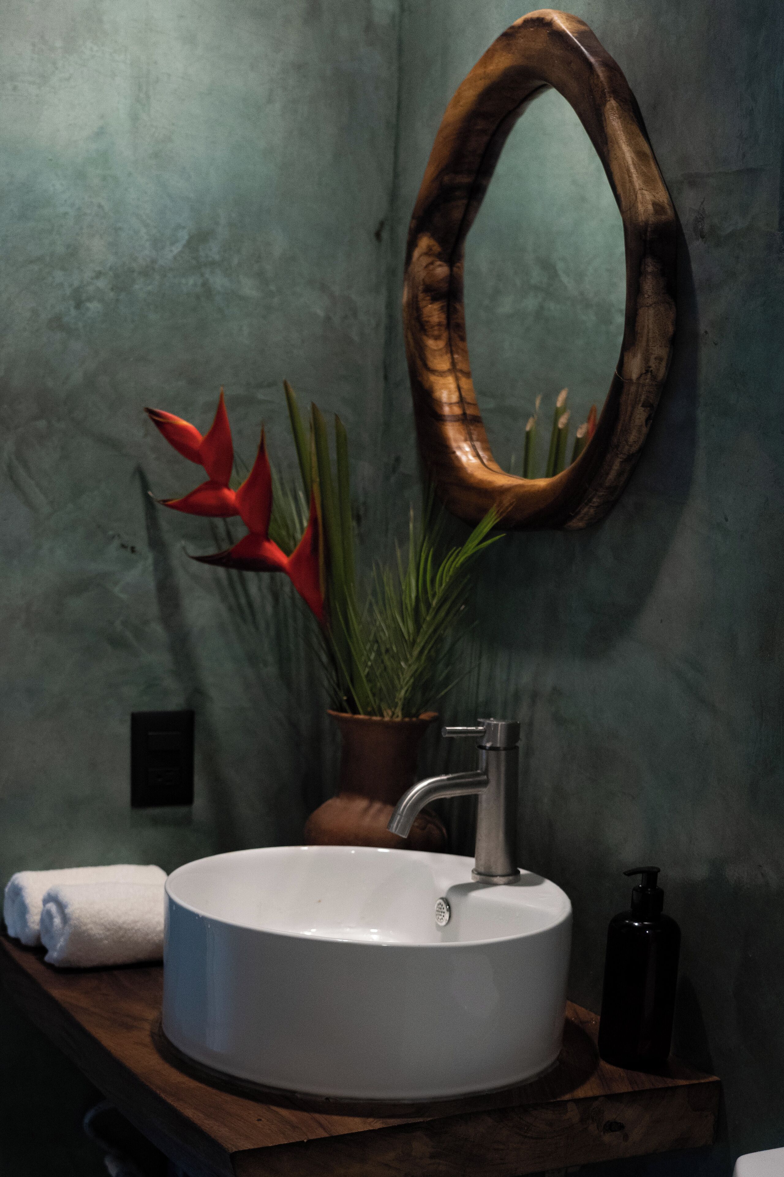 Un baño con un lavabo redondo, un espejo con marco de madera y una planta en maceta con flores rojas.