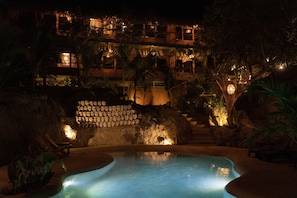 Vista nocturna de un complejo turístico con piscina, muros de piedra y zonas de estar al aire libre.