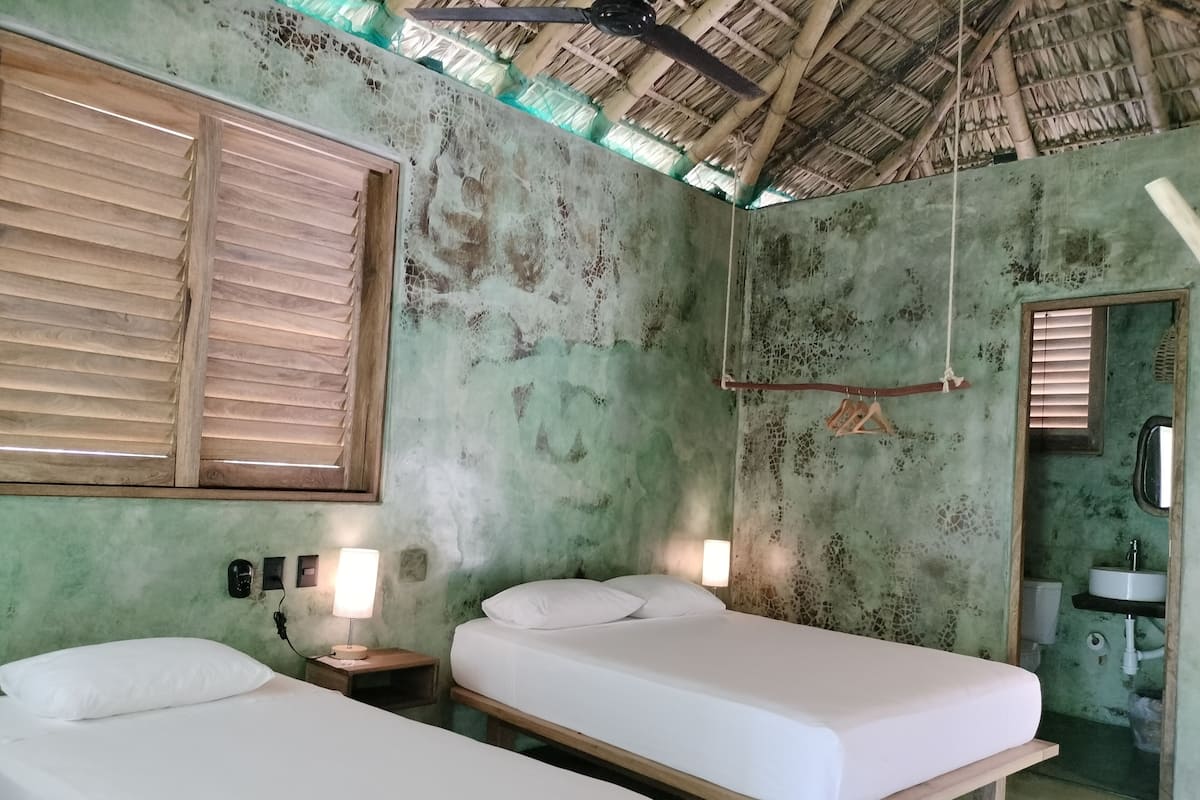 Ein Zimmer mit zwei Einzelbetten, Holzboden, einem Fenster mit Holzjalousien, einem Nachttisch mit Lampe und einem Badezimmer mit Waschbecken und Spiegel.