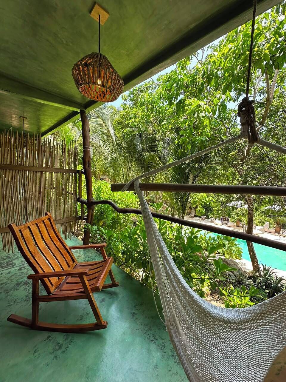 A hammock chair and a wooden rocking chair on a porch with a view of a pool and lush greenery.