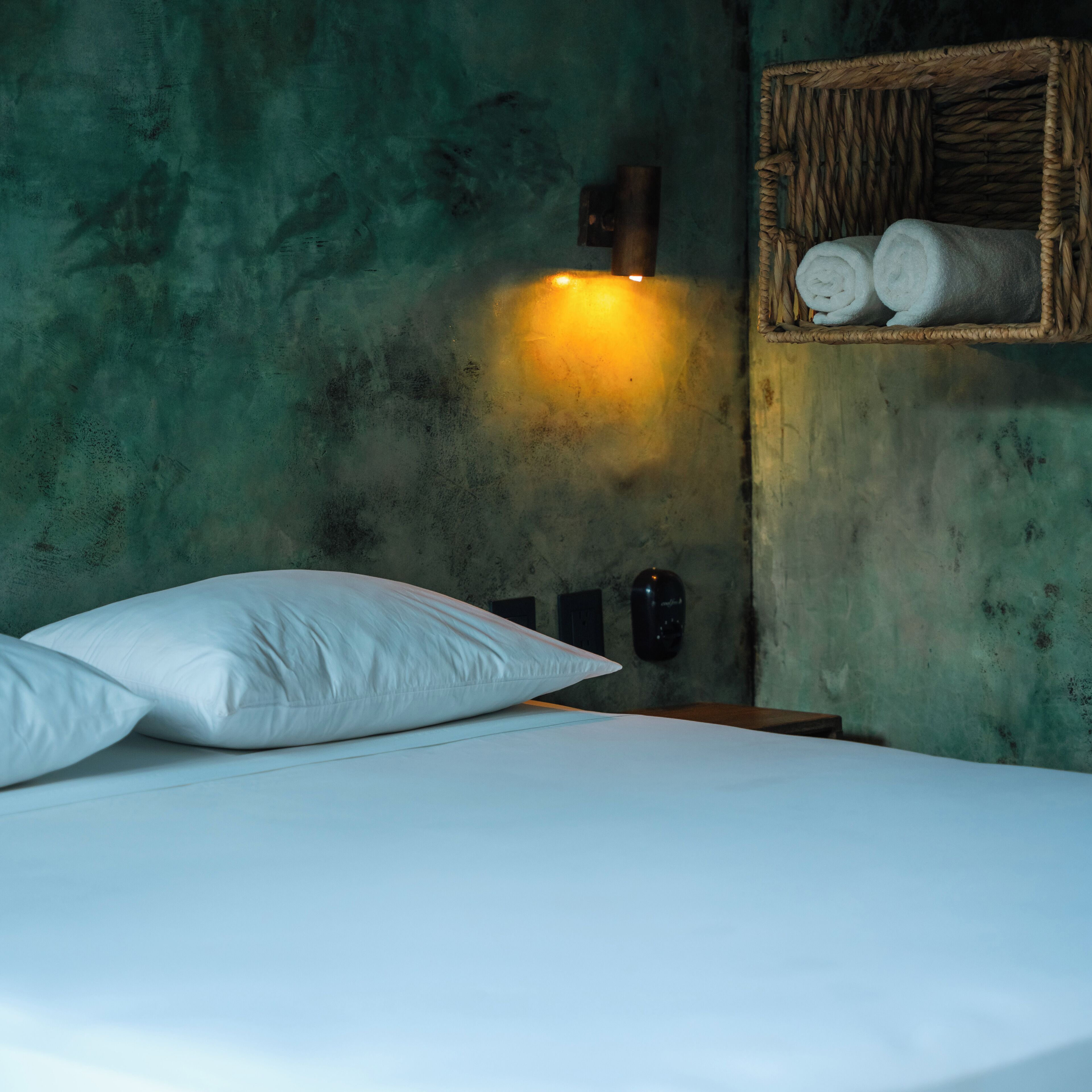 Una cama con almohadas blancas y cabecera frente a una pared verde texturizada. Se ve una lámpara de pared y una cesta de mimbre con toallas enrolladas.