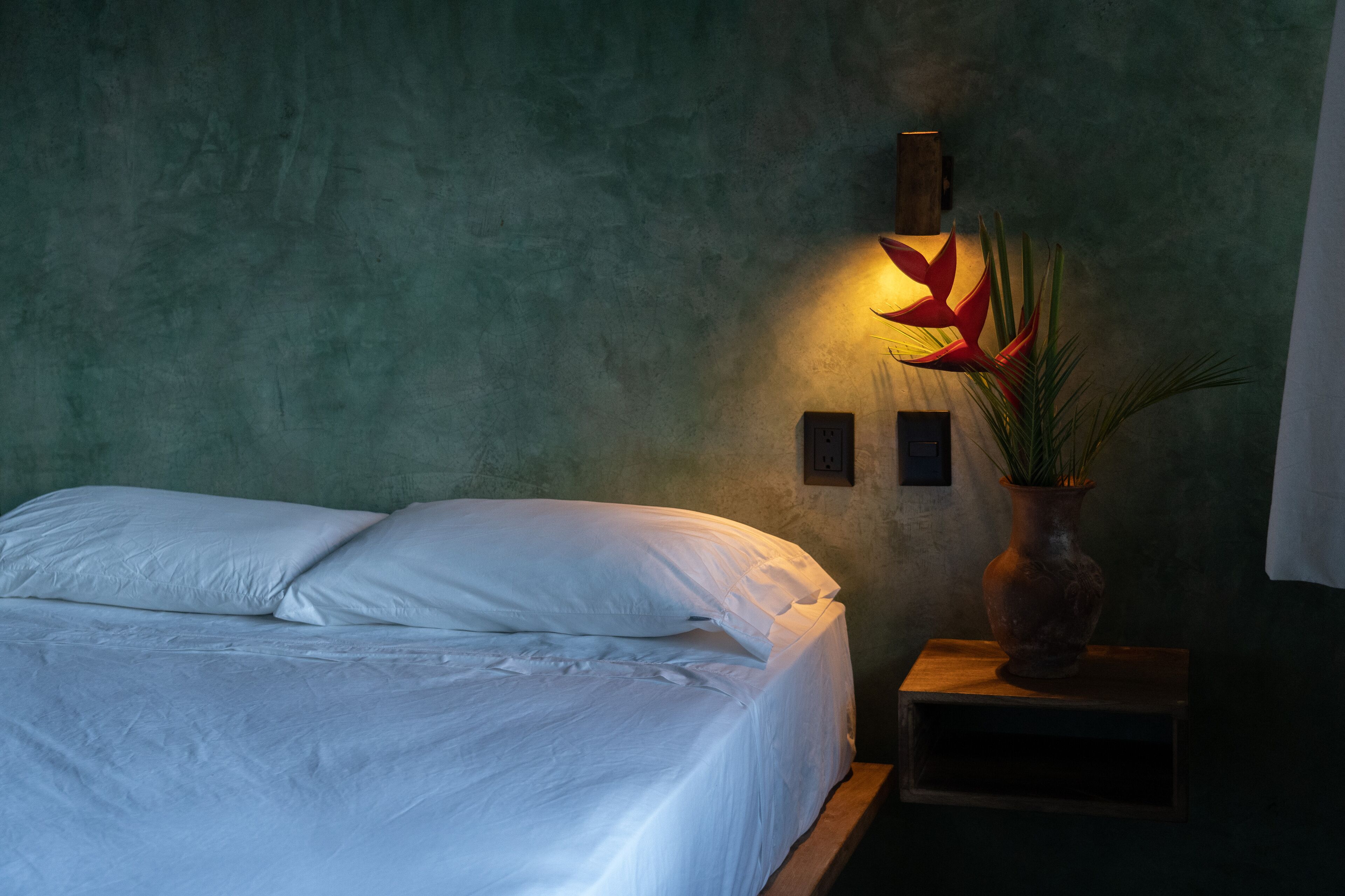 A neatly made bed with white linens in a room with a textured green wall. There is a wall-mounted light fixture with a red flower design and a potted plant on a small wooden nightstand.