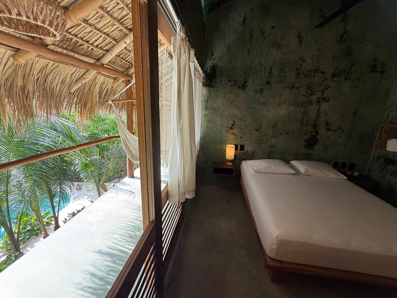 Una habitación con cama, mesita de noche, lámpara, ventana con cortinas y vista a una piscina y palmeras.