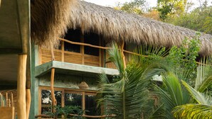 A two-story building with a thatched roof, surrounded by lush greenery and a sandy ground. - Cocos Hotel (San Pedro Pochutla)