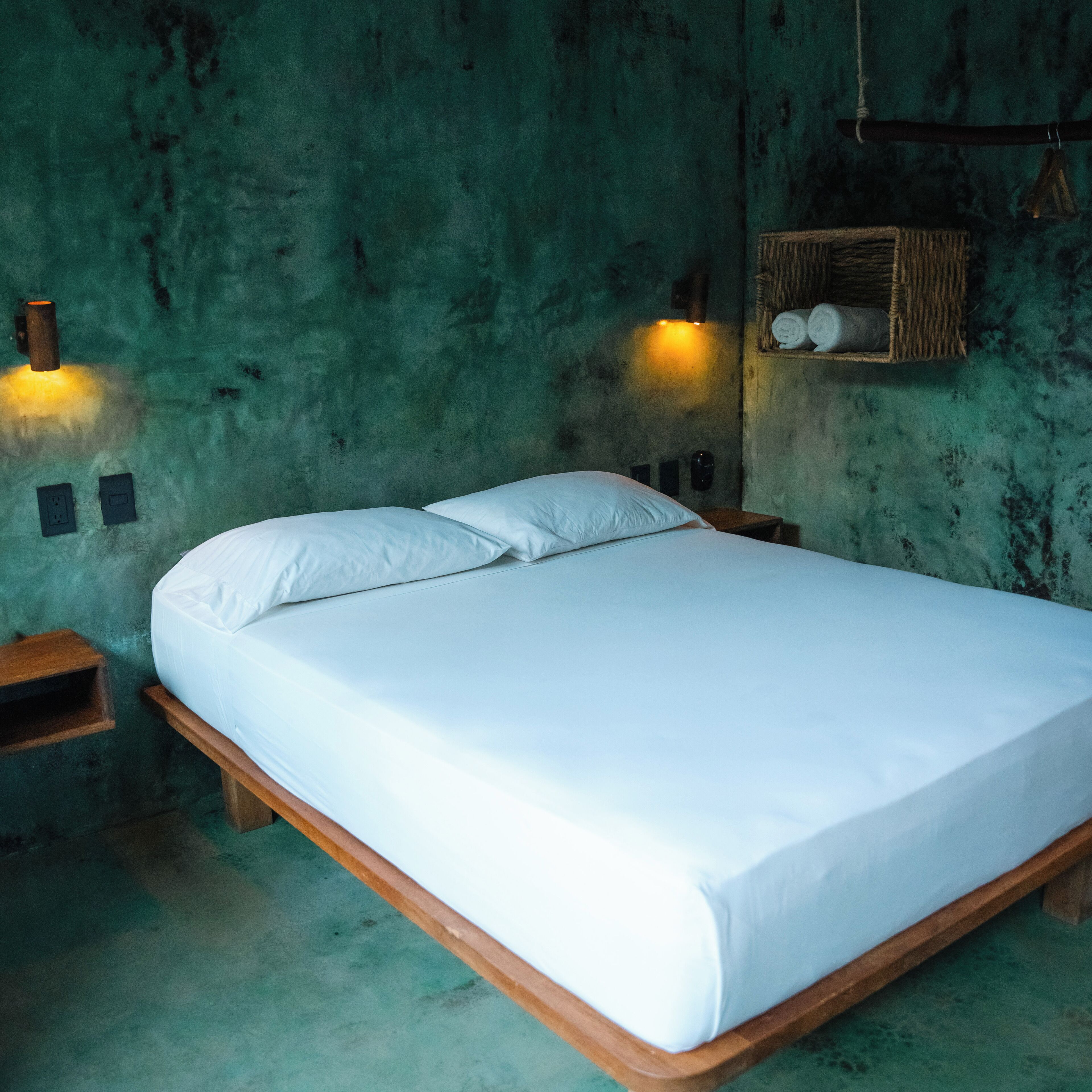 A single bed with white linens in a room with green walls and a rustic wooden floor. There are two wall-mounted lights and a small shelf with rolled towels.