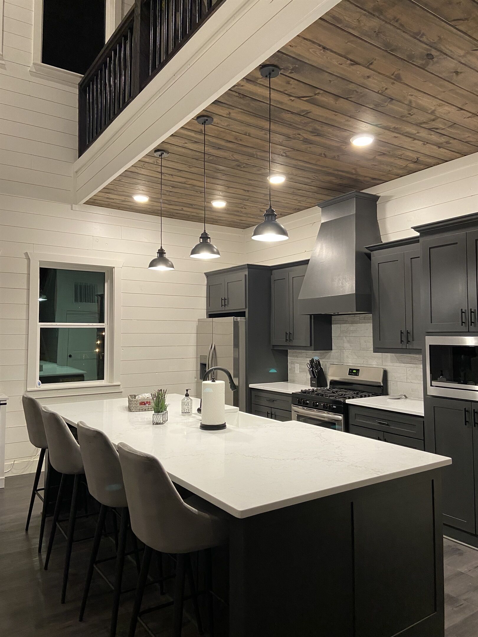 Une cuisine moderne dotée d’un îlot central, de meubles de rangement aux couleurs sombres et de luminaires suspendus.