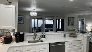 A modern kitchen with white cabinetry, a built-in dishwasher, a coffee station, and a large window with curtains.