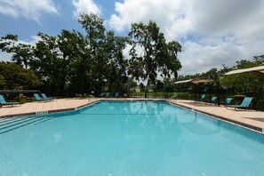 Ein rechteckiger Pool mit gefliestem Rand, umgeben von Liegestühlen und Sonnenschirmen, mit Bäumen und blauem Himmel im Hintergrund.