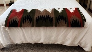 A neatly made bed with a white comforter and a decorative blanket featuring geometric patterns. There are two bedside tables, each with a lamp, and a small blue object on one of the tables. - Charming 3 bedroom Ranch in scenic McElmo Canyon (Cortez)