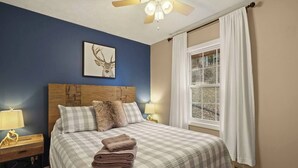 A bedroom with a wooden headboard, a large window with white curtains, a checkered bedspread, and a folded towel on the bed.
